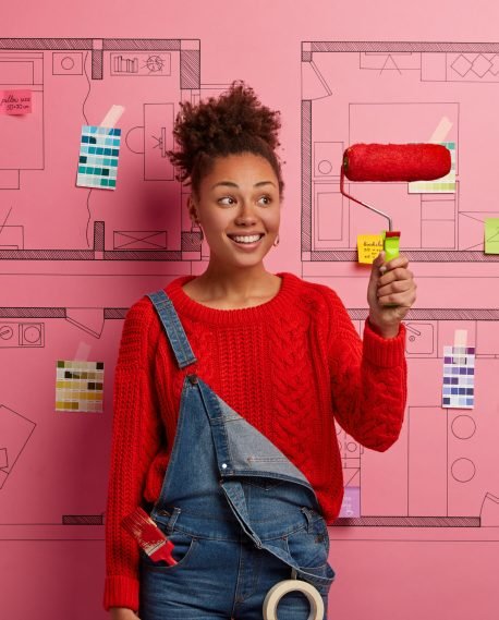 Glad smiling Afro American decorator holds paint roller, busy doing repairing according to design project, paints room red, wears knitted sweater and denim overalls, uses tools for repair and painting